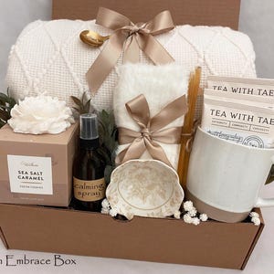 May include: A gift box containing a white textured throw blanket with a gold ribbon, a white mug, three Tea With Tae packets, a Sea Salt Caramel candle, a brown spray bottle labeled Calming Spray, a small floral bowl, and a gold spoon.