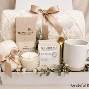 May include: A gift box containing a cream-colored knit blanket with a tan ribbon, a Moonstruck milk chocolate bar, a white candle in a textured glass, Tea with Tae sachets, a gold spoon, and a white ceramic mug with a tan base.