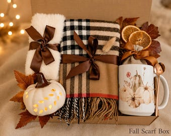 May include: A gift box containing a white fuzzy scarf, a tan and black plaid scarf with fringe, a white ceramic pumpkin with cutouts, a white mug with a pumpkin print, a copper spoon, dried orange slices, and brown ribbon. Text reads Fall Scarf Box.