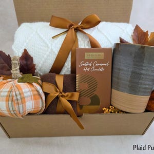 May include: A gift box containing a white knit blanket tied with a bronze ribbon, a plaid fabric pumpkin, a brown box of Beshaler Salted Caramel Hot Chocolate, and a two-toned gray and tan ceramic mug, all accented with artificial autumn leaves.