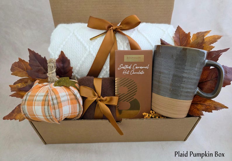 May include: A gift box containing a white knit blanket tied with a bronze ribbon, a plaid fabric pumpkin, a brown box of Beshaler Salted Caramel Hot Chocolate, and a two-toned gray and tan ceramic mug, all accented with artificial autumn leaves.