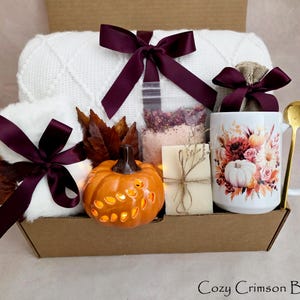 May include: A gift box containing a white knit blanket with a dark purple ribbon, a white mug with a floral and pumpkin print, a gold spoon, a small orange ceramic pumpkin, a bar of soap, a bag of bath salts, and faux autumn leaves.
