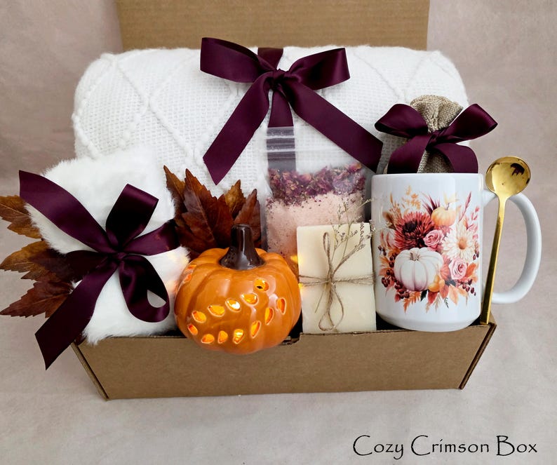 May include: A gift box containing a white knit blanket with a dark purple ribbon, a white mug with a floral and pumpkin print, a gold spoon, a small orange ceramic pumpkin, a bar of soap, a bag of bath salts, and faux autumn leaves.