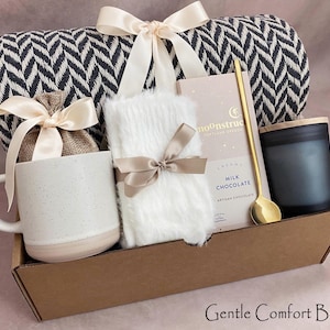 May include: A gift box containing a black and white patterned blanket, a white mug, a white fluffy item, a candle, a chocolate bar, and a gold spoon. The items are arranged in a brown cardboard box and tied with ribbons. The text "Gentle Comfort Box" is at the bottom.
