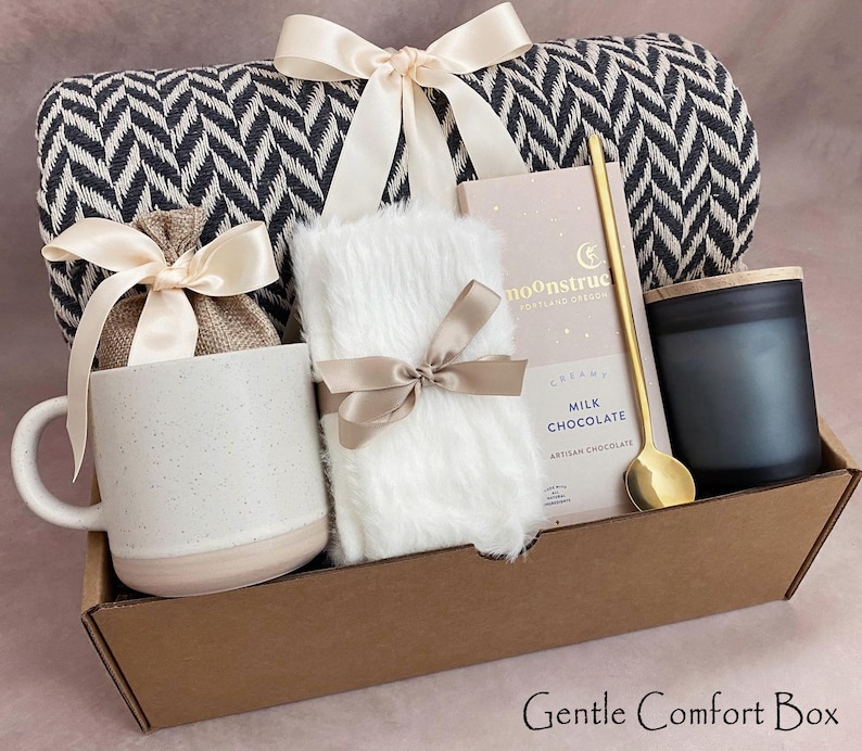 May include: A gift box containing a black and white patterned blanket, a white mug, a white fluffy item, a candle, a chocolate bar, and a gold spoon. The items are arranged in a brown cardboard box and tied with ribbons. The text "Gentle Comfort Box" is at the bottom.