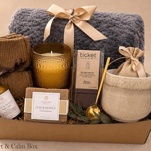 May include: A gift box containing a grey textured throw blanket with a beige ribbon, a pair of brown knit socks, a gold candle, a bar of Oat & Honey soap, a Ticket Chocolate bar, a gold spoon, a beige mug, and a small hessian pouch.