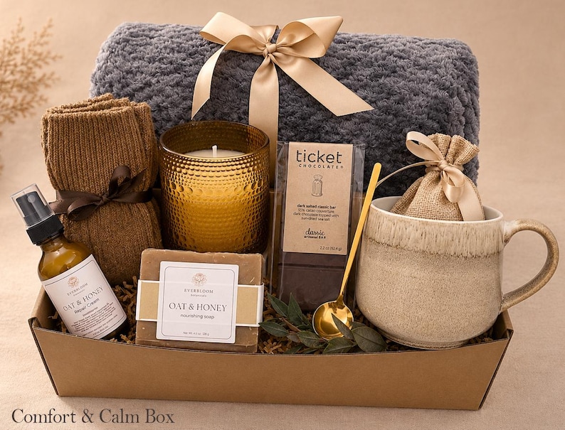 May include: A gift box containing a grey textured throw blanket with a beige ribbon, a pair of brown knit socks, a gold candle, a bar of Oat & Honey soap, a Ticket Chocolate bar, a gold spoon, a beige mug, and a small hessian pouch.