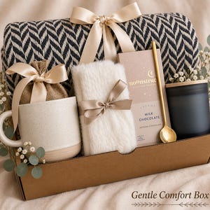 May include: A gift box containing a black and white herringbone throw blanket with a beige ribbon, a speckled ceramic mug, a burlap pouch, a white faux fur item, a bar of Noonstruck milk chocolate, a gold spoon, and a matte black candle.