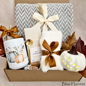May include: A gift box containing a blue and white herringbone throw blanket with a cream ribbon, a white mug with pumpkin illustrations, a gold spoon, a milk chocolate bar, white faux fur fabric, and a white ceramic pumpkin with leaf cutouts.