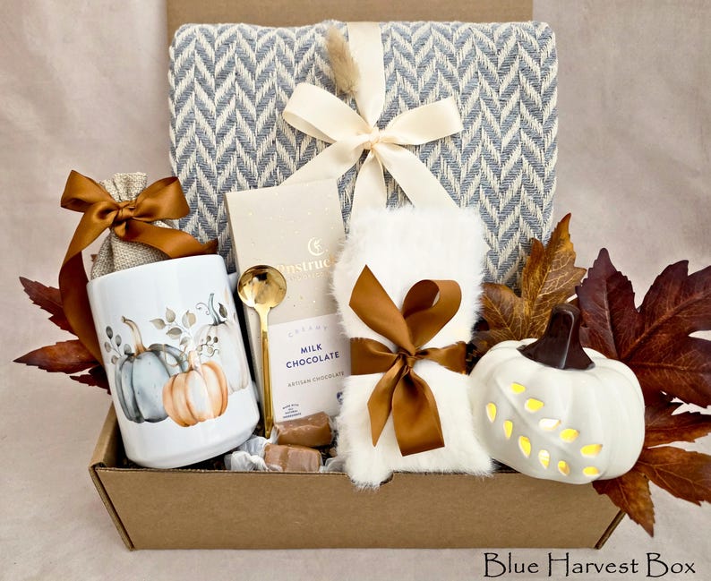 May include: A gift box containing a blue and white herringbone throw blanket with a cream ribbon, a white mug with pumpkin illustrations, a gold spoon, a milk chocolate bar, white faux fur fabric, and a white ceramic pumpkin with leaf cutouts.