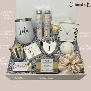 May include: A gift box filled with self-care items. Includes a white tumbler with "Lola" printed on it, bath salts, a rhinestone necklace, an oatmeal bath bomb, a scrunchie, and other items. The box is lined with white paper and has a light pink background.