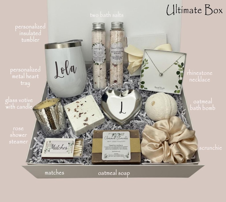 May include: A gift box filled with self-care items. Includes a white tumbler with "Lola" printed on it, bath salts, a rhinestone necklace, an oatmeal bath bomb, a scrunchie, and other items. The box is lined with white paper and has a light pink background.