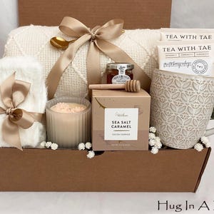 May include: A gift box containing a white blanket, a patterned mug, tea bags, a candle, honey, and cookies. The box is tied with a tan ribbon. The mug has a floral design. The box is brown and has the text "Hug In A Box".