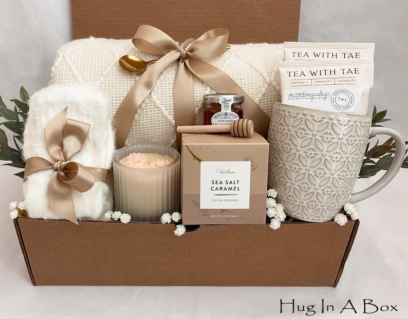 May include: A gift box containing a white blanket, a patterned mug, tea bags, a candle, honey, and cookies. The box is tied with a tan ribbon. The mug has a floral design. The box is brown and has the text "Hug In A Box".