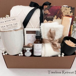 May include: A brown gift basket containing self-care items. It features a white mug with tea bags, a white blanket, a candle, honey, and Oat Honey products. The basket is tied with a black and a tan bow. The text "Timeless Retreat Box" is at the bottom.