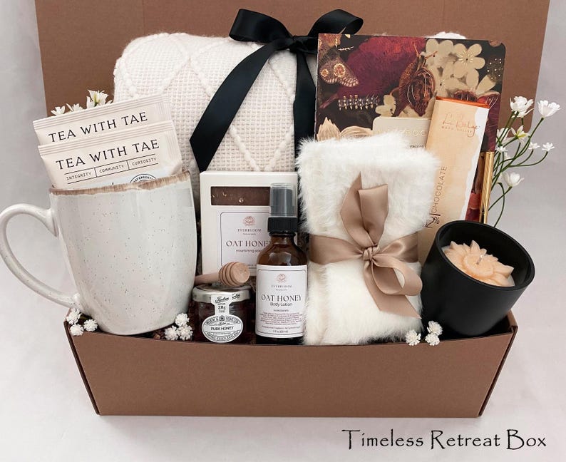 May include: A brown gift basket containing self-care items. It features a white mug with tea bags, a white blanket, a candle, honey, and Oat Honey products. The basket is tied with a black and a tan bow. The text "Timeless Retreat Box" is at the bottom.