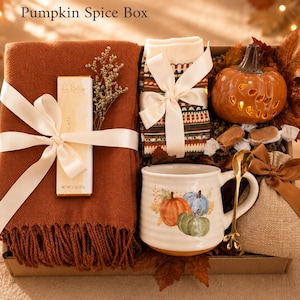 May include: A gift box labeled Pumpkin Spice Box containing a rust-colored fringed throw blanket, a chocolate bar, patterned socks, a ceramic mug with pumpkin illustrations, a gold-toned spoon, a small burlap pouch, and a ceramic pumpkin lantern.