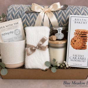 May include: A gift box containing a blue and white chevron throw blanket with a cream ribbon, a speckled ceramic mug, a packet of Tea With Tae, a white fuzzy item with a brown ribbon, a jar of honey, a wooden dipper, and Rustic Bakery shortbread.