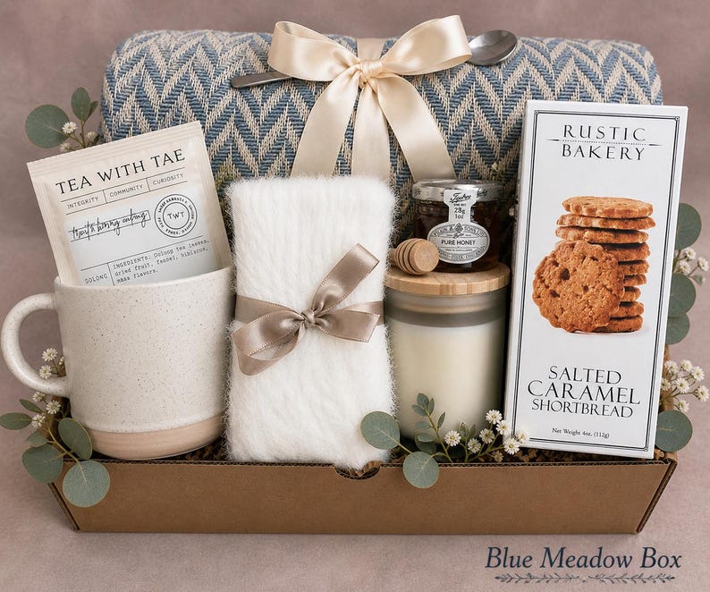 May include: A gift box containing a blue and white chevron throw blanket with a cream ribbon, a speckled ceramic mug, a packet of Tea With Tae, a white fuzzy item with a brown ribbon, a jar of honey, a wooden dipper, and Rustic Bakery shortbread.