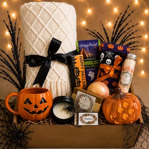 May include: A Halloween gift box containing a cream-colored knit throw with a black ribbon, an orange pumpkin mug, a black candle with a spider, a box of matches, a bar of soap, a bath bomb, a bottle of bath salts, chocolate, and pumpkin decor.