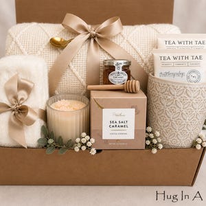 Puede incluir: Una caja de regalo que contiene una manta de punto color crema con una cinta beige, un art&iacute;culo blanco y esponjoso con cinta beige, una vela de vidrio acanalado, un tarro de miel con cuchara de madera, galletas de caramelo y una taza estampada.