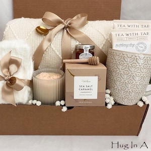 May include: A gift box filled with a white blanket, a mug, tea packets, a candle, honey, and cookies. The box is tied with a tan ribbon. The mug has a floral pattern. The box is brown and has the text "Hug In A Box".