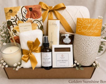 May include: A gift basket containing a floral notebook, a white fuzzy item with a gold ribbon, a candle, a bottle of Oat Honey body lotion, a bar of Oat Honey soap, a chocolate bar, a jar of honey with a dipper, and a textured mug with a card.
