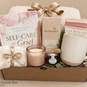 May include: A gift box containing a book titled Self-Care for Grief, a Moonstruck milk chocolate bar, a white ceramic mug, three Tea With Tae packets, a pink ribbed glass candle, a small jar of honey with a dipper, and a white faux fur pouch.