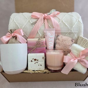 May include: A gift box containing self-care products. It features a white mug with a pink base, a candle, soap, bath salts, a washcloth, and rose-scented lotion. The items are arranged in a brown box and tied with pink ribbons. The text "Blush Box" is at the bottom.