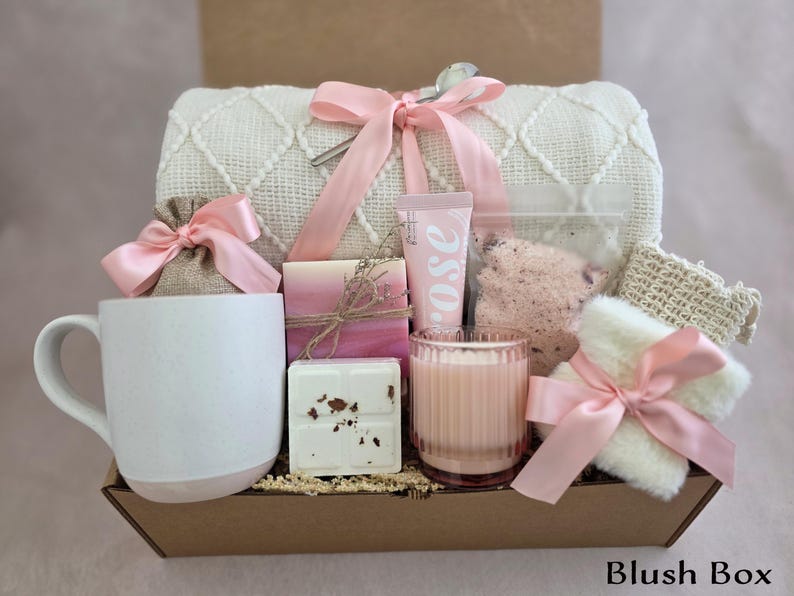 May include: A gift box containing self-care products. It features a white mug with a pink base, a candle, soap, bath salts, a washcloth, and rose-scented lotion. The items are arranged in a brown box and tied with pink ribbons. The text "Blush Box" is at the bottom.