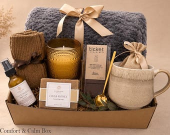 May include: A gift box containing a gray textured throw blanket with a beige ribbon, a pair of brown knit socks, a gold candle, a bar of Oat & Honey soap, a Ticket Chocolate bar, a gold spoon, a beige mug, and a small burlap pouch.