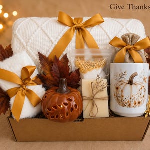 May include: A Give Thanks Box gift set containing a white cable-knit blanket with a gold ribbon, a white mug with a pumpkin print, a gold spoon, a ceramic pumpkin, a bar of soap, a small burlap bag, and faux autumn leaves in a cardboard box.