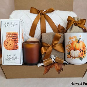 May include: A gift box containing a white knit blanket with a brown ribbon, a box of Rustic Bakery Salted Caramel Shortbread, a brown candle, a tan cloth bundle with a brown ribbon, two caramels, and a white mug with pumpkins and a gold spoon.