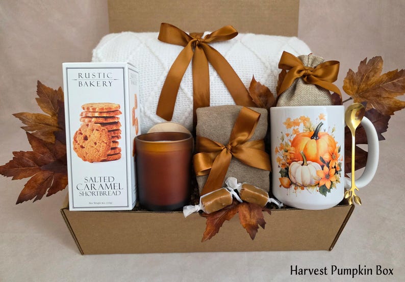 May include: A gift box containing a white knit blanket with a brown ribbon, a box of Rustic Bakery Salted Caramel Shortbread, a brown candle, a tan cloth bundle with a brown ribbon, two caramels, and a white mug with pumpkins and a gold spoon.
