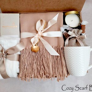 May include: A gift box containing a beige fringed scarf tied with a cream ribbon and a gold spoon, a white textured mug, a white faux fur item with a beige ribbon, a candle in a gold tin, and a small burlap pouch with the text Moonstruck.