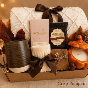 May include: A gift basket containing a cream-colored knit throw with a brown ribbon, a dark brown ceramic mug with a gold spoon, a Moonstruck chocolate bar, a Sorrento mocha mix packet, cream-colored socks, a candle in an orange pumpkin jar, and leaves.