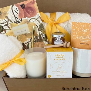 May include: A sunshine-themed gift box filled with items like a candle, tea, cookies, honey, and a white towel. The box is decorated with a yellow ribbon and the words "Sunshine Box" are printed on the bottom.