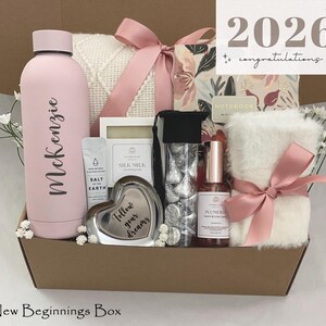 May include: A gift box filled with a pink water bottle, a white blanket, a heart-shaped trinket, and other items. The box is tied with a pink ribbon and includes the text "2026 congratulations". The water bottle has the name "McKenzie" on it.