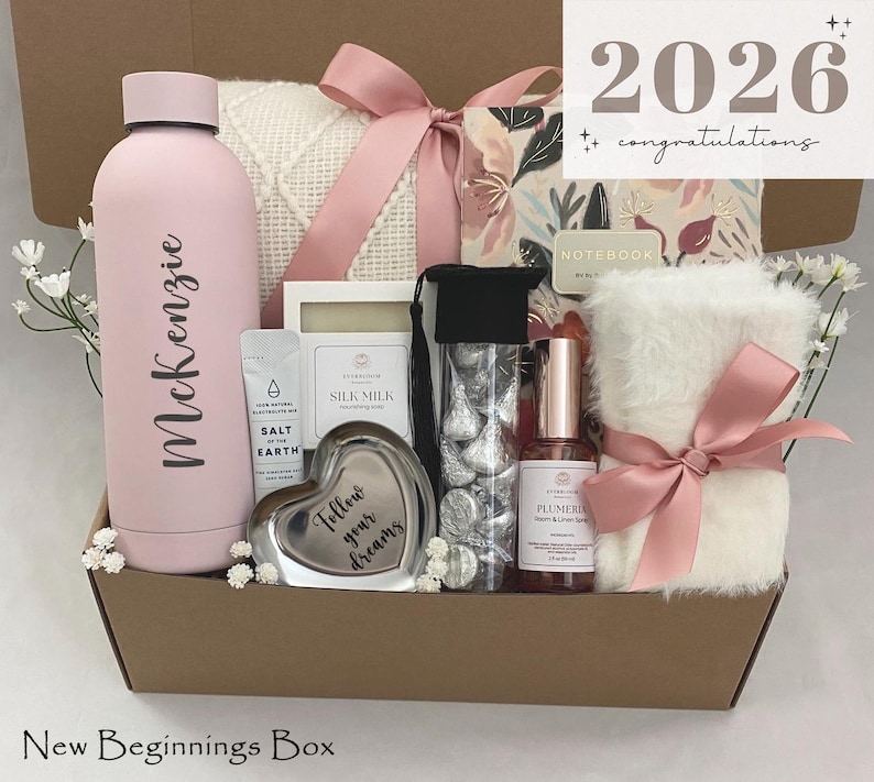 May include: A gift box filled with a pink water bottle, a white blanket, a heart-shaped trinket, and other items. The box is tied with a pink ribbon and includes the text "2026 congratulations". The water bottle has the name "McKenzie" on it.