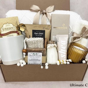 May include: A gift box containing self-care items such as a white mug with caramel mocha, a gold spoon, a white blanket, a candle, shea lotion, and other spa products. The box is tied with a beige ribbon. The text "Ultimate Cozy Box" is at the bottom.