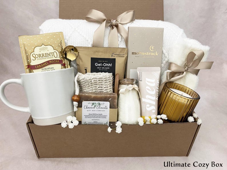 May include: A gift box containing self-care items such as a white mug with caramel mocha, a gold spoon, a white blanket, a candle, shea lotion, and other spa products. The box is tied with a beige ribbon. The text "Ultimate Cozy Box" is at the bottom.