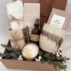 May include: A gift box containing self-care products. It features a white mug, chocolate bars, a candle, a bottle of calming spray, a fluffy white item tied with a ribbon, and a bar of soap. The box is decorated with greenery and the text "Still Moments Box".