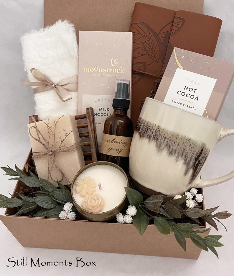 May include: A gift box containing self-care products. It features a white mug, chocolate bars, a candle, a bottle of calming spray, a fluffy white item tied with a ribbon, and a bar of soap. The box is decorated with greenery and the text "Still Moments Box".
