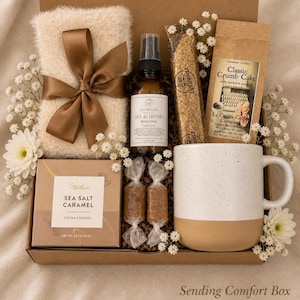 May include: A gift box containing a fuzzy cream-coloured cloth with a brown ribbon, a bottle of Oat & Honey body lotion, a package of Classic Crumb Cake coffee, a box of Sea Salt Caramel cocoa cookies, two wrapped sweets, and a speckled ceramic mug.