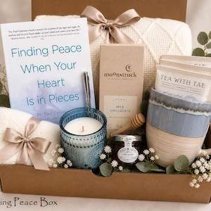 May include: A gift box filled with self-care items. It includes a book titled "Finding Peace When Your Heart Is in Pieces", a blue candle, a mug, chocolate, honey, tea, and a blanket. The box is tied with a beige ribbon.
