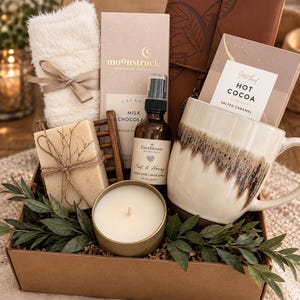 May include: A gift box containing a cream-colored fuzzy pair of socks with a ribbon, a Moonstruck milk chocolate bar, a brown leather-bound journal, a packet of salted caramel hot cocoa, a bar of soap, a room spray, and a candle.