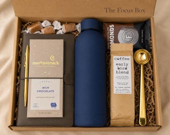 May include: A gift box labeled The Focus Box containing a dark blue water bottle, a gold pen, a notebook, a Moonstruck milk chocolate bar, a bag of Early Bird Blend coffee, a gold coffee scoop, and a package of dark chocolate almonds.