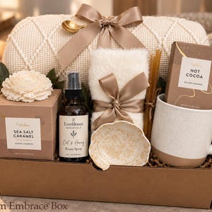 May include: A gift box containing a cream-colored knit blanket with a satin bow, a box of Sea Salt Caramel cocoa cookies, an Everbloom Botanicals Oat & Honey room spray, a white faux fur item, a small patterned dish, and a Hot Cocoa mug.