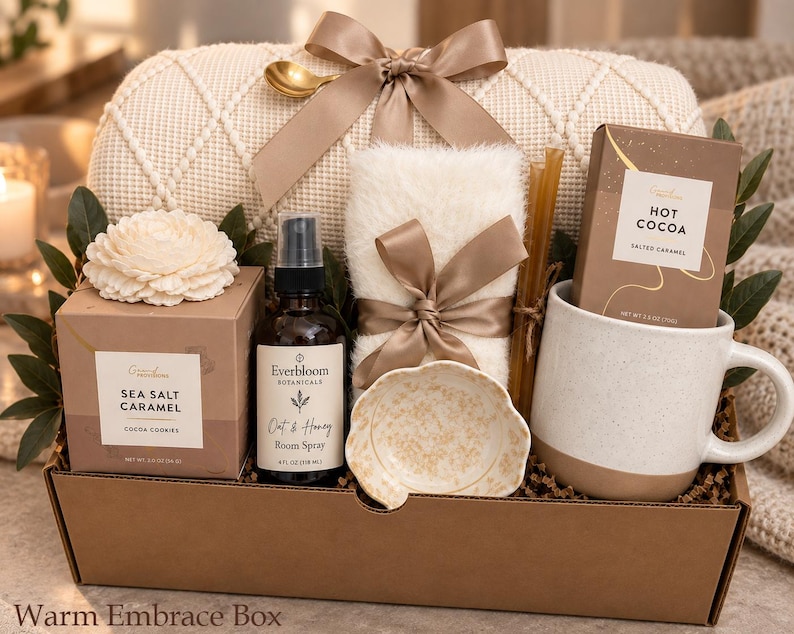 May include: A gift box containing a cream-colored knit blanket with a satin bow, a box of Sea Salt Caramel cocoa cookies, an Everbloom Botanicals Oat & Honey room spray, a white faux fur item, a small patterned dish, and a Hot Cocoa mug.