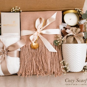 May include: A gift box containing a dusty rose fringed scarf tied with a cream ribbon and gold spoon, a white faux fur item with a satin ribbon, a white textured mug, a candle in a gold tin, a small burlap bag, and a box labeled moonstruck.